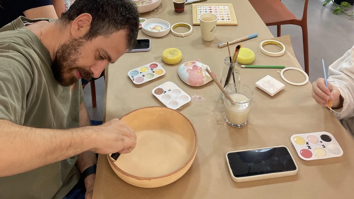 אבא צובע קערת קרמיקה ליד שולחן עם צבעים ומכחולים, וילדה צובעת לידו