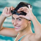 Portrait of fit young woman wearing swimming cap and goggles at the pool. Portrait of a smiling female swimmer wearing swim goggles at swimming pool. Happy fit girl at pool looking at camera.