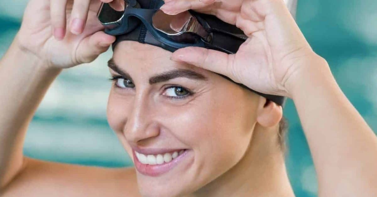 Portrait of fit young woman wearing swimming cap and goggles at the pool. Portrait of a smiling female swimmer wearing swim goggles at swimming pool. Happy fit girl at pool looking at camera.